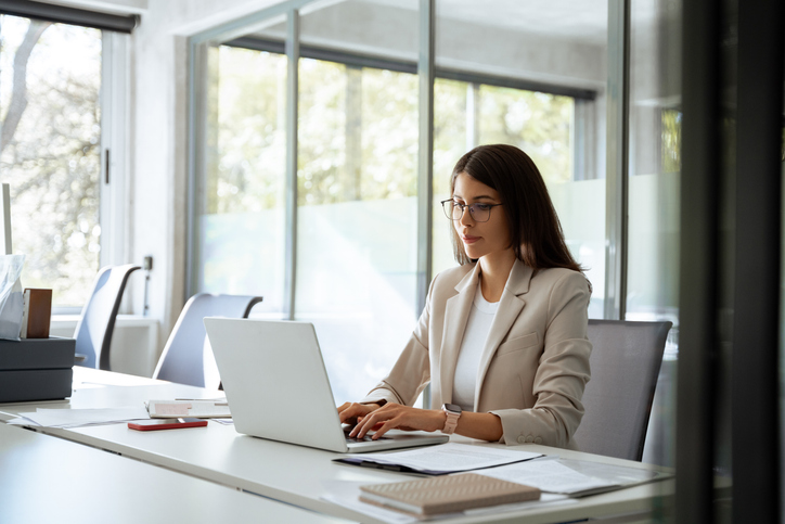 Professional woman working on a laptop at a modern office desk with documents and natural light from large windows.