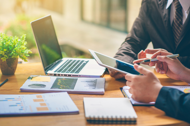 Business professionals reviewing financial charts on a tablet and laptop at a desk during a work meeting.