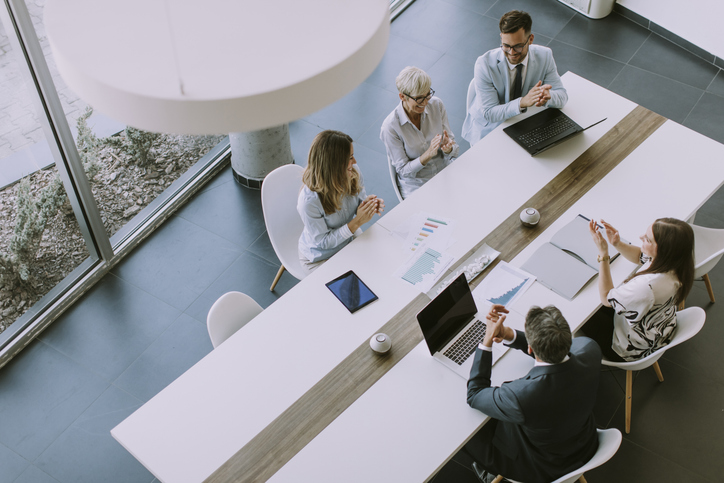 Business team seated around a conference table during a meeting, viewed from above, with laptops and documents on the table.