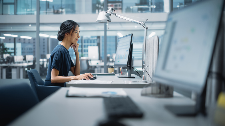 Woman working at a computer workstation in a modern office, focused on data displayed on her monitor.
