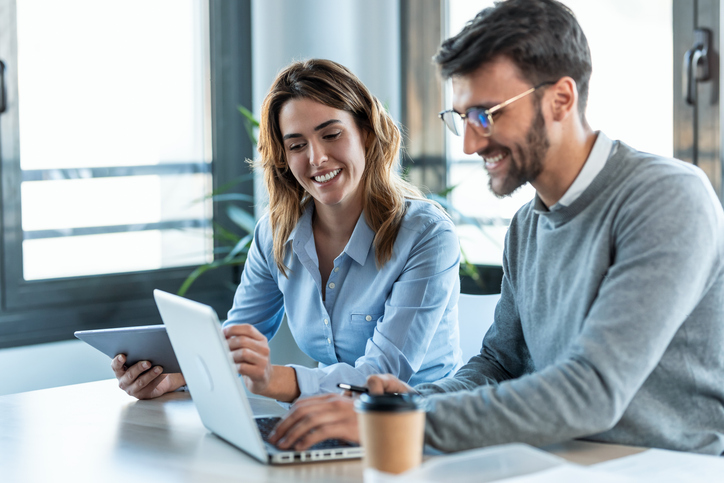 Two coworkers collaborating at a desk while reviewing content on a laptop and tablet in a bright office.