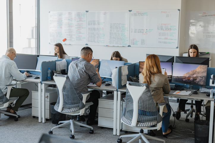 Employees working at computer stations in an open office, with multiple monitors and a whiteboard covered in notes behind them.