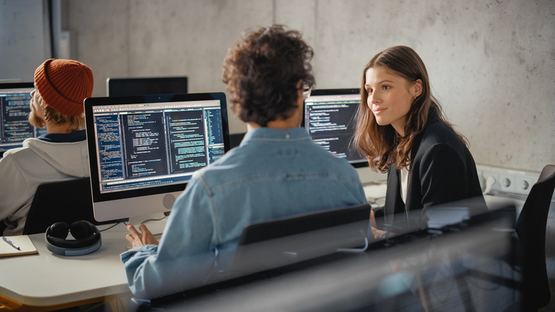 Software developers working together at computer stations with code displayed on multiple monitors in a modern office.