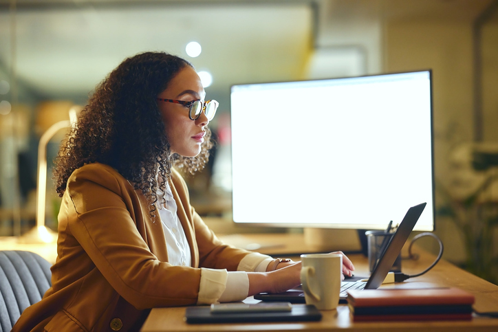 Woman working on a laptop at her desk in a softly lit office, focused on her screen with a coffee nearby.