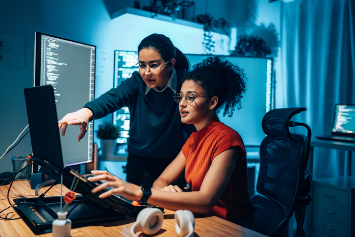 Two women working together at a computer workstation, reviewing code displayed on multiple monitors in a modern office.