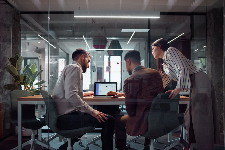 Three coworkers collaborating around a laptop in a glass meeting room within a modern office.