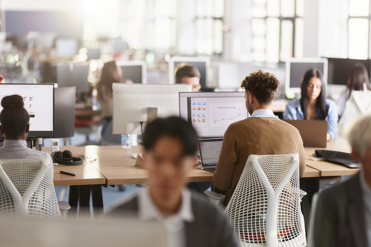 Employees working at computer desks in a bright open office with multiple monitors and laptops.