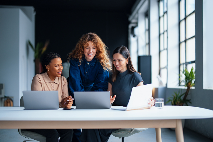 Three coworkers collaborating at a table while working on laptops in a bright, modern office.