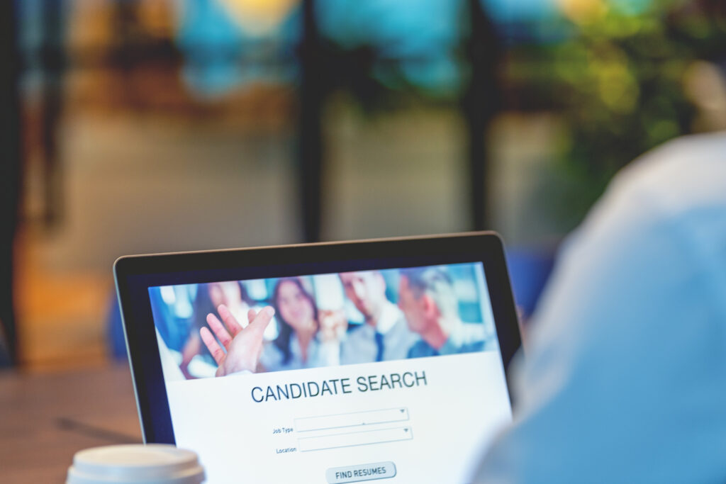 laptop screen displaying a "candidate search" interface for recruiting, with a hand reaching out to the viewer.